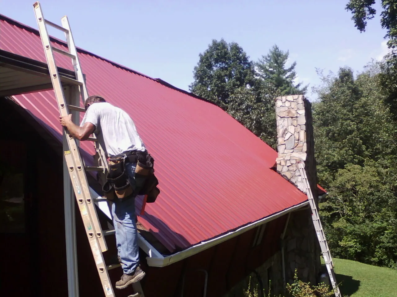 Roofer on a ladder beside a red metal roof and stone chimney — Soffit & Fascia Repair by Roofing Brothers in Creighton